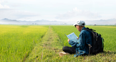 Fototapeta na wymiar Asian boy in plaid shirt wears cap and has a backpack, holding a map and a binoculars, sitting on ridge rice field of asian farmers to study landscapes, mountains, rice paddy field, bird watching.