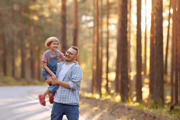 Fototapeta premium A magical moment unfolds as a father holds little daughter in his arms and they laugh joyously while strolling along a forest road, surrounded by majestic pine trees and the warm glow of the sunset.