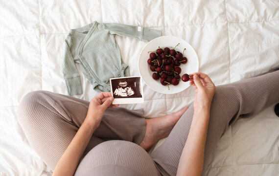 High angle top view of unrecognizable woman sitting on bed looking at ultrasound photo eating black berries at cozy home. Expecting baby in summer or spring concept.