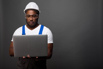 Serious black master in protective helmet looks at laptop. Dark-skinned foreman concentrates on printing on laptop a report on completed construction and commissioning of facility. 