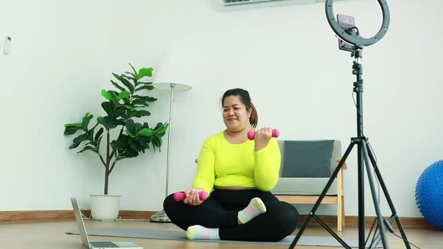Asian fat woman streamer sits exercising lifting dumbbells study from trainer on tablet internet online with live streaming video recording on smartphone using ring light brighten up during broadcast.
