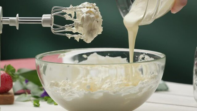 Pouring melted white chocolate into glass bowl with whipped cream; hand mixer with tasty cream on twisters; homemade strawberry dessert