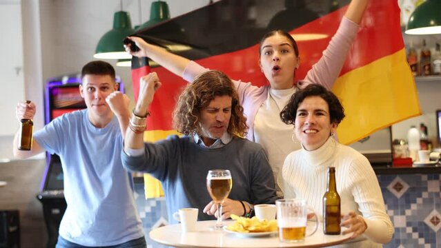 Group of young adult sports fans rooting for favorite team and waving flag of Germany while watching match together in pub
