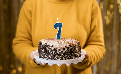 Woman holding birthday cake with burning candles number 7 against decorated background, close up....