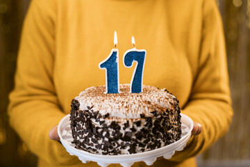 Woman holding birthday cake with burning candles number 17 against decorated background, close up. Celebrates birthday holiday at home.