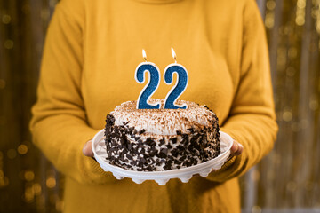 Woman holding birthday cake with burning candles number 22 against decorated background, close up....