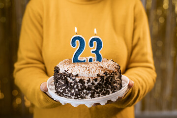 Woman holding birthday cake with burning candles number 23 against decorated background, close up....