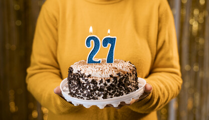 Woman holding birthday cake with burning candles number 27 against decorated background, close up....