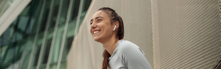 Smiling woman in sportswear resting after run in city leaning against the wall and holding phone