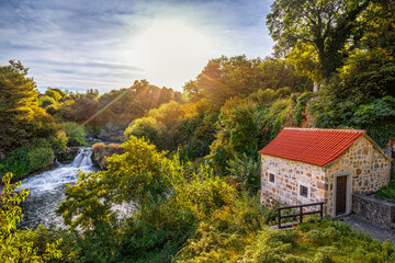 Krka, Croatia - Beautiful summer sunrise scenery of small stone house at Krka Waterfalls in Krka National Park, Dalmatia with warm morning colored foliage, rising sun and blue sky