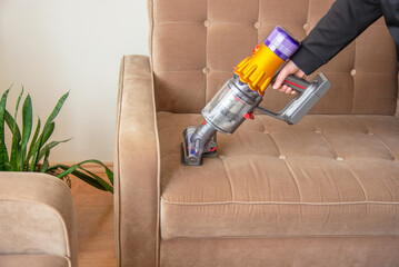 Young woman vacuums sofa with cordless handheld vacuum cleaner. House cleaning.