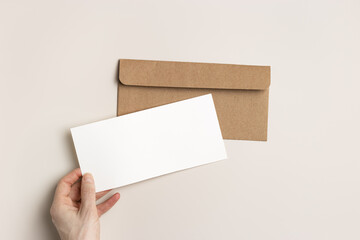 Blank paper card mock up in woman hand, craft envelope on beige background. White invitation on table. Open paper letter mockup. Minimal modern lifestyle photo. Flat lay, top view, copy space.