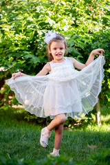 A cheerful little girl plays in the park on the lawn in a beautiful dress rejoices in the summer. Beautiful white girl smiling while walking in the courtyard of the house.