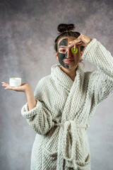 Young woman wear white bathrobe with holding kiwi fruit with black mask on her face
