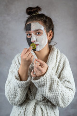 Young lady in white bathrobe applying white and black clay masks on face and holding cucumber slice