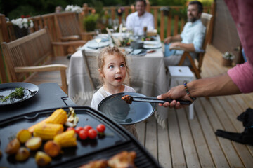 Father putting grilled meat and vegetable on plate to his daughter during family summer garden party.