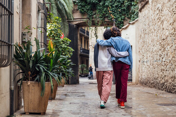Fototapeta na wymiar rear view of a gay male couple walking embraced along a beautiful ancient street decorated with plants, concept of leisure and love between people of the same sex