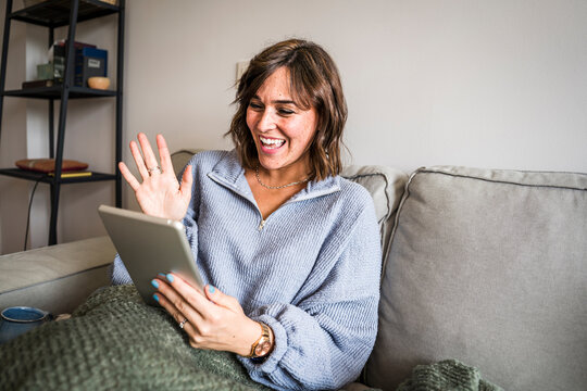 Happy woman on video call waving through tablet PC on sofa at home