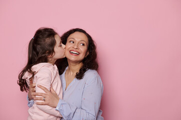 Caucasian cute little girl kissing her mother who smiles and looks aside at copy ad space....