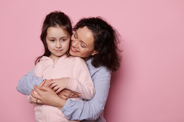 Happy pretty woman hugging her little kid daughter from behind, isolated on pink background studio portrait. Mommy and child have fun together. Mother's Day. Love Family Parenthood Childhood concept