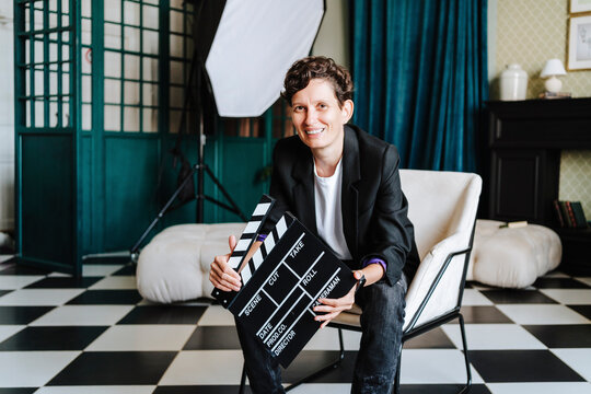 Smiling director sitting on chair holding clapboard at film set