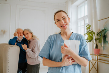 Smiling nurse holding tablet PC with senior man and woman in background at home