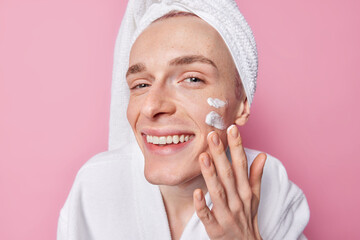 Close up shot of cheerful man with freckled skin applies facial cream for skin moisturising wears...