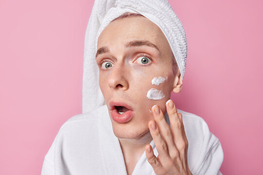 Headshot of freckled adult man applies facial cream after showerring wears white bathrobe and towel wrapped on head isolated over pink background tries to moisturise skin. Daily skincare concept