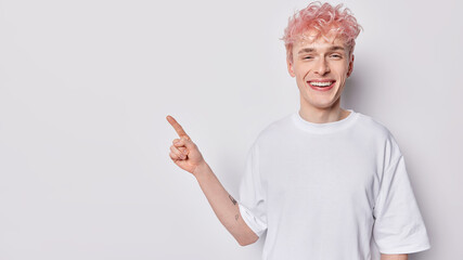 Young pink haired man points index finger aside on blank space invites or advertises promo deal...