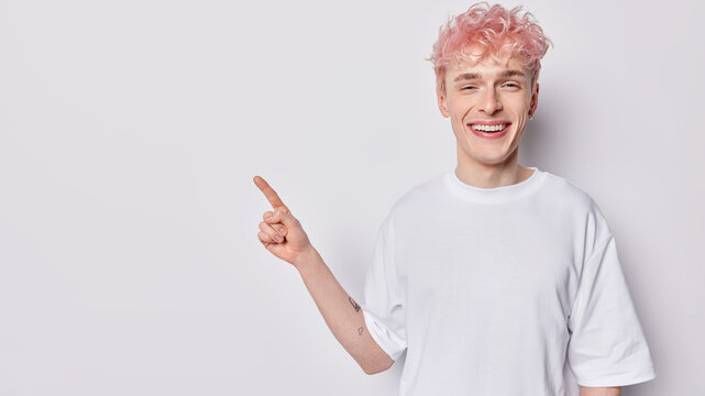 Young pink haired man points index finger aside on blank space invites or advertises promo deal stands against white background dressed in basic t shirt smiles gladfully. Place your text here