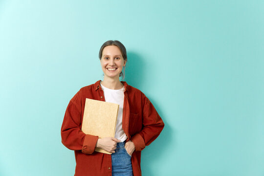 Smiling woman with hand in pocket holding file near teal wall