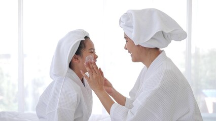 Asian mother adding treatment cream on the cheek to little girl with spa dress and head covered...