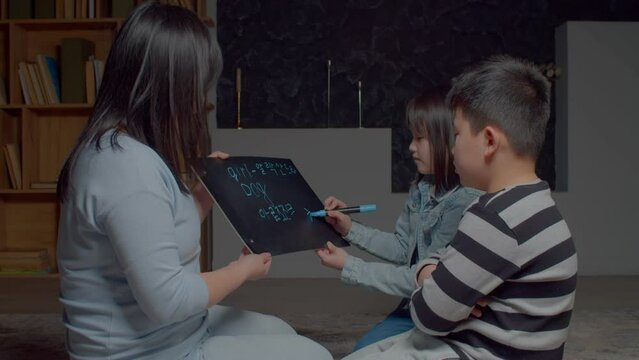 Positive adorable elementary age Asian children with loving attractive mother learning Korean language, reading and pronouncing words , written on portable chalkboard during homeschooling.