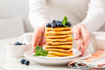 Morning, woman hands serving homemade breakfast, stack of pancakes with blueberries and honey or maple syrup. White table background.