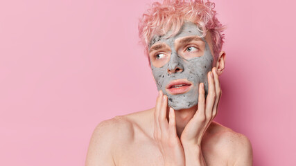 Horizontal shot of thoughtful man keeps hands on cheeks focused aside attentively applies facial nourishing clay mask stands shirtless against pink background copy space for your advetising content