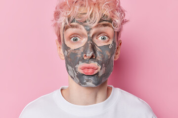 Close up portrait of surprised man with pink hair looks wondered at camera pouts lips wears beauty clay mask applies moisturizer on skin dressed in white t shirt poses against rosy studio background