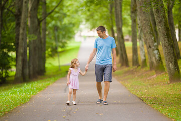 Little daughter and young adult father speaking and walking on sidewalk through tree alley at city park. Spending time together in beautiful warm sunny summer day. Front view.