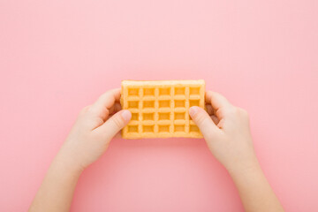 Little child hands holding fresh golden waffle on light pink table background. Pastel color....