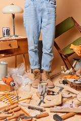 Cropped shot of unrecognizable carpenter dressed in denim overalls boots surrounded by tools uses lots of equipment works in carpentry workshop reconstructs old wooden furniture. Profession concept