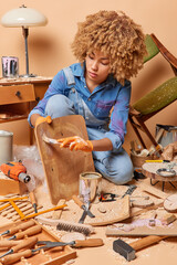 Vertical shot of curly haired female carpenter paints wooden surface with paint renews or renovates...