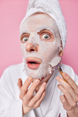 Close up shot of shocked man enjoys moisturizing procedure applies beauty sheet mask uses facial massager stares impressed keeps mouth opened wears white bathrobe and towel on head pink background