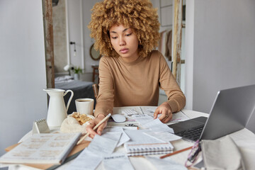 Photo of serious woman looks through paper documents and receipts manages business affairs plans future investments or household budget poses at desktop with laptop computer works from home.