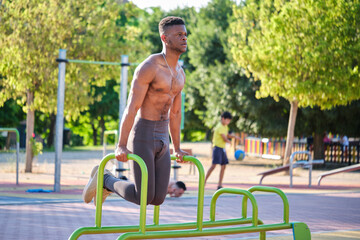 Young fit shirtless black man doing calisthenics workout on parallel bars outdoors on sunny day....
