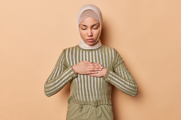 Calm Arabian Muslim woman wears traditional hijab presses hands on chest makes grateful gesture poses with eyes closed isolated over brown studio background. People and religious lifestyle concept