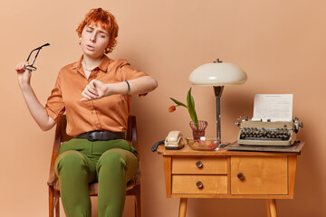 Serious elegant vintage lady poses on chair waits for business partner holds spectacles in hand checks time on watch sits on old chair near table with stationary phone typewriter and retro lamp
