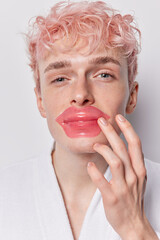 Close up shot of serious pink haired man applies collagen moisturizing mask patch on lips undergoes beauty routine at home wears domestic robe isolated over white background. Korean cosmetics concept