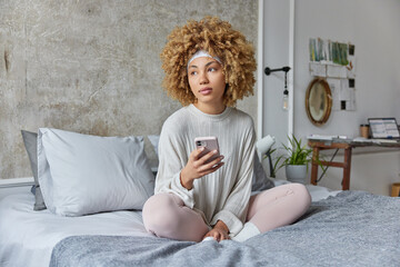 Pensive curly haired woman dressed in comfortable pajama checks mails or social networks on...
