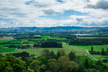 Fototapeta na wymiar Aerial view over lush green farmland, distand mountain range rising over horizon under cloudy sky. Breathtaking New Zealand Landscape. Greys Hill Lookout, Gisborne, North Island, New Zealand