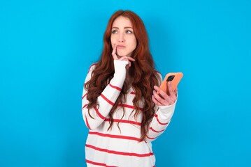 young woman wearing striped T-shirt over blue background thinks deeply about something, uses modern...