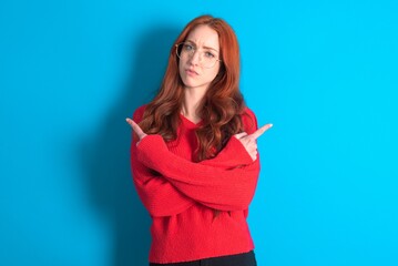 Serious young woman wearing red sweater over blue background crosses hands and points at different sides hesitates between two items. Hard decision concept